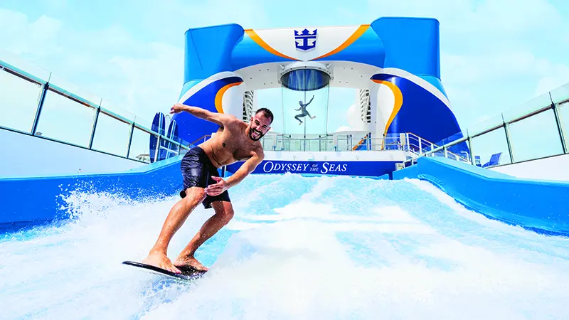 A guest is using the FlowRider surf simulator while another guest is sky diving in RipCord by iFLY in the background on Royal Caribbean’s Odyssey of the Seas.
