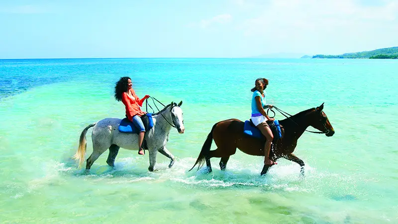 Two women riding horses through the water in Jamaica