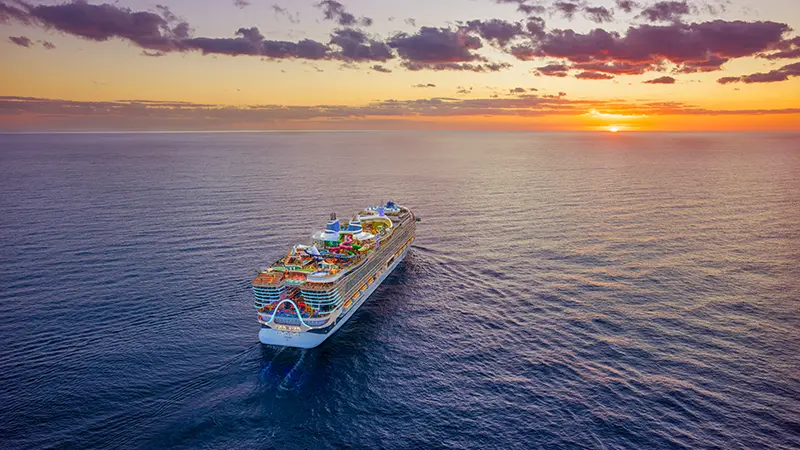 A Royal Caribbean cruise ship sailing through a calm ocean at sunset.